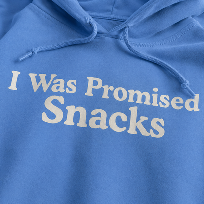 A close-up of a blue hoodie with the phrase 'I Was Promised Snacks' printed in large cream-colored letters across the chest.