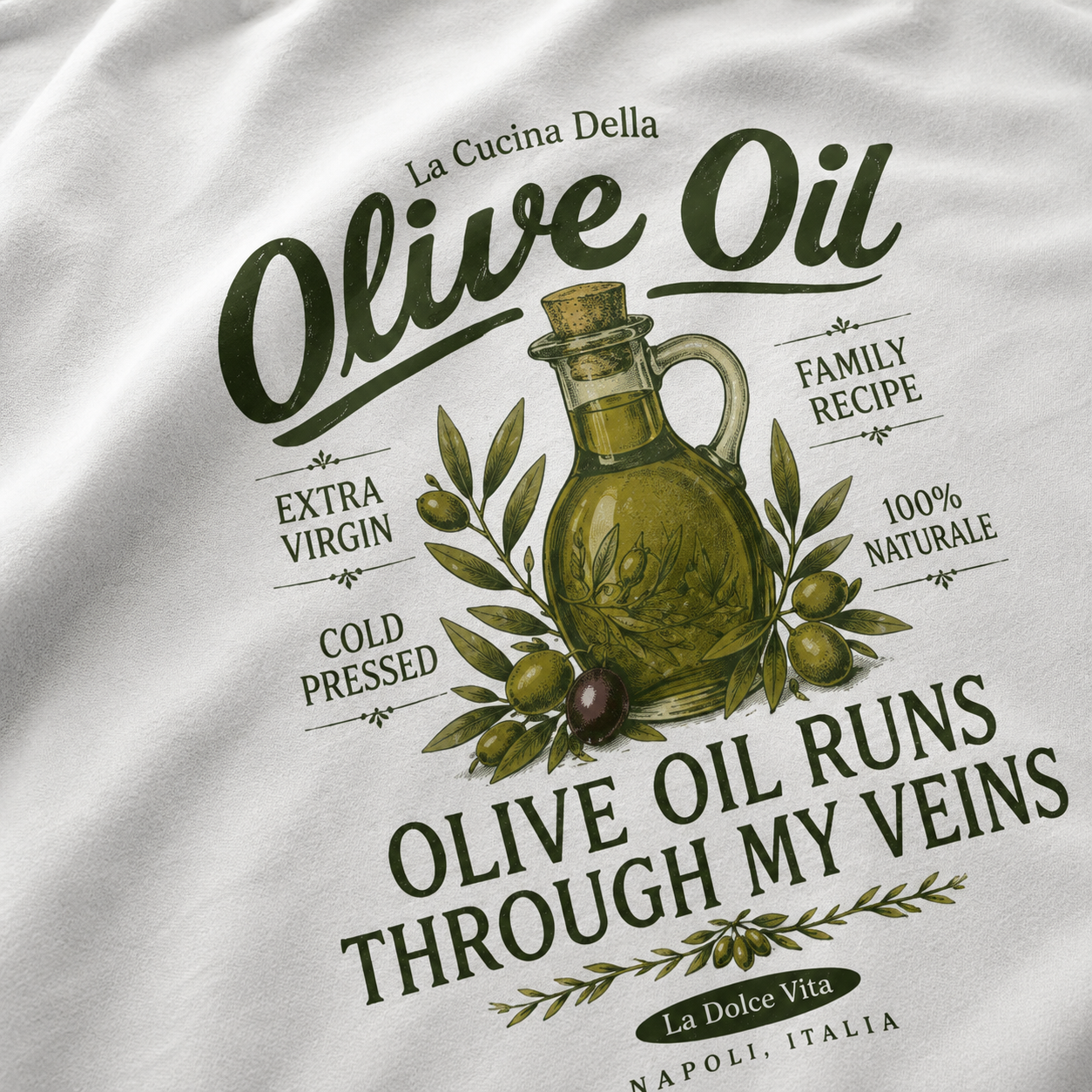 A close-up of a white t-shirt featuring a detailed olive oil graphic print. The design includes a vintage-style olive oil jug surrounded by olives and leaves, with Italian text such as 'La Cucina Della', 'Olive Oil', 'Extra Virgin', 'Cold Pressed', 'Family Recipe', '100% Naturale', and 'Olive Oil Runs Through My Veins'.