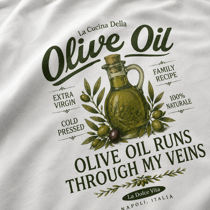 A close-up of a white t-shirt featuring a detailed olive oil graphic print. The design includes a vintage-style olive oil jug surrounded by olives and leaves, with Italian text such as 'La Cucina Della', 'Olive Oil', 'Extra Virgin', 'Cold Pressed', 'Family Recipe', '100% Naturale', and 'Olive Oil Runs Through My Veins'.