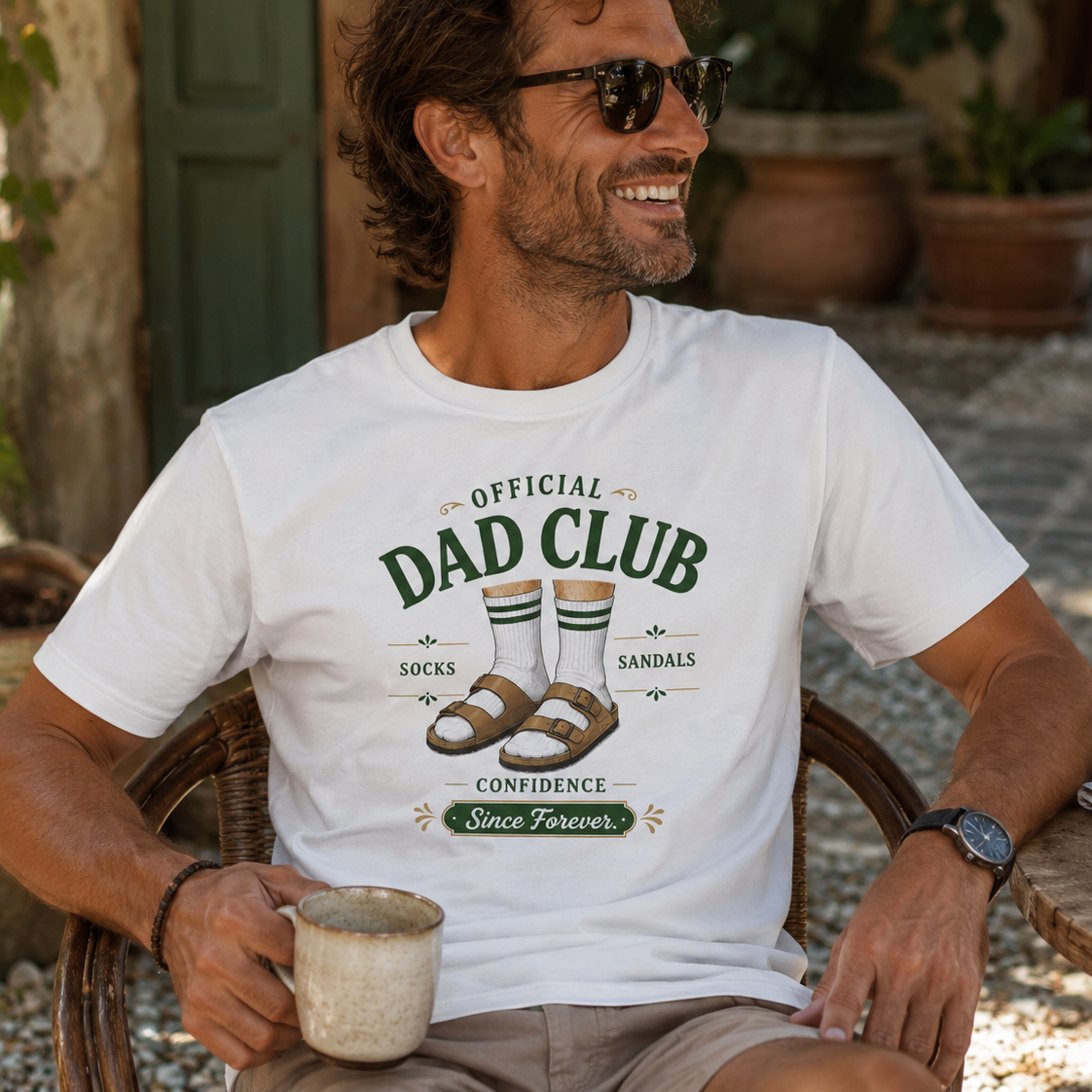 A man wearing a white graphic t-shirt that reads 'OFFICIAL DAD CLUB' with an illustration of socks, sandals, and the words 'CONFIDENCE Since Forever'. He is sitting outdoors in a wicker chair holding a mug.
