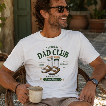 A man wearing a white graphic t-shirt that reads 'OFFICIAL DAD CLUB' with an illustration of socks, sandals, and the words 'CONFIDENCE Since Forever'. He is sitting outdoors in a wicker chair holding a mug.