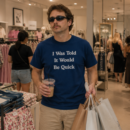 A man wearing a navy blue graphic t-shirt with the white text 'I Was Told It Would Be Quick' walks through a clothing store while holding shopping bags and an iced coffee.
