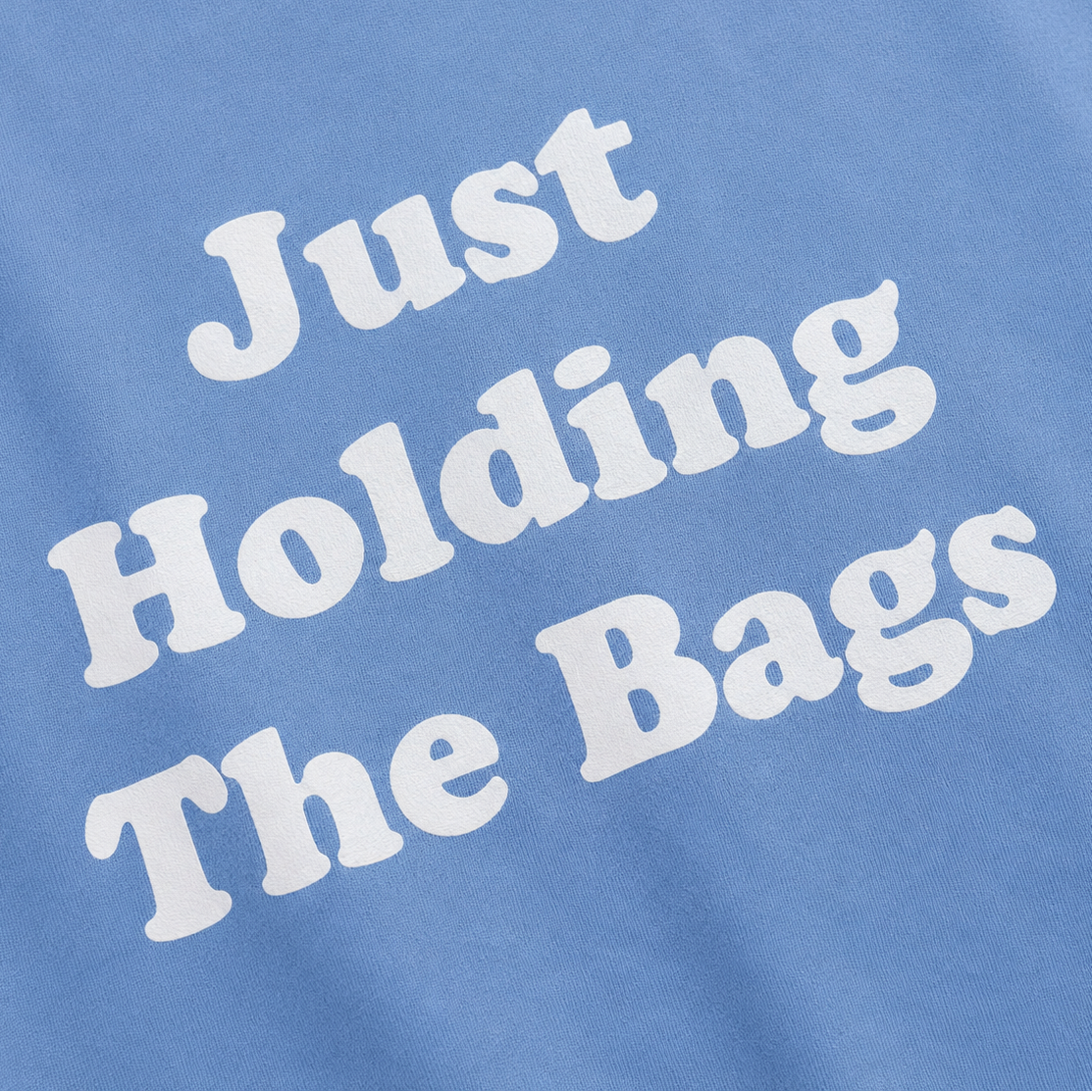 Close-up of a light blue t-shirt with the phrase 'Just Holding The Bags' printed in a bold, white, retro-style font.