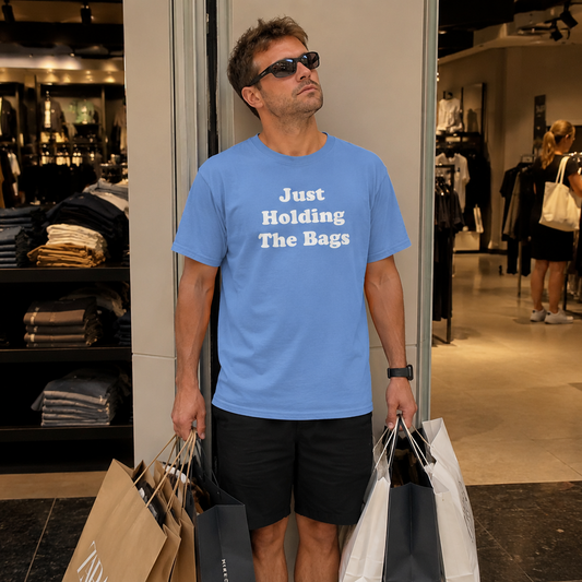 A man wearing sunglasses and a light blue graphic t-shirt with the text 'Just Holding The Bags' stands in a clothing store holding shopping bags.