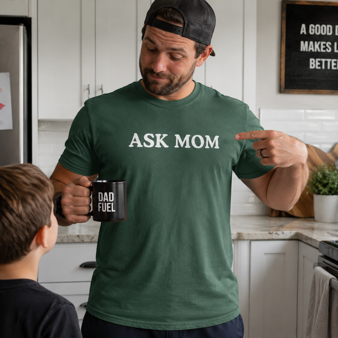 Man in a kitchen wearing a green t-shirt with 'Ask Mom' text, holding a mug.