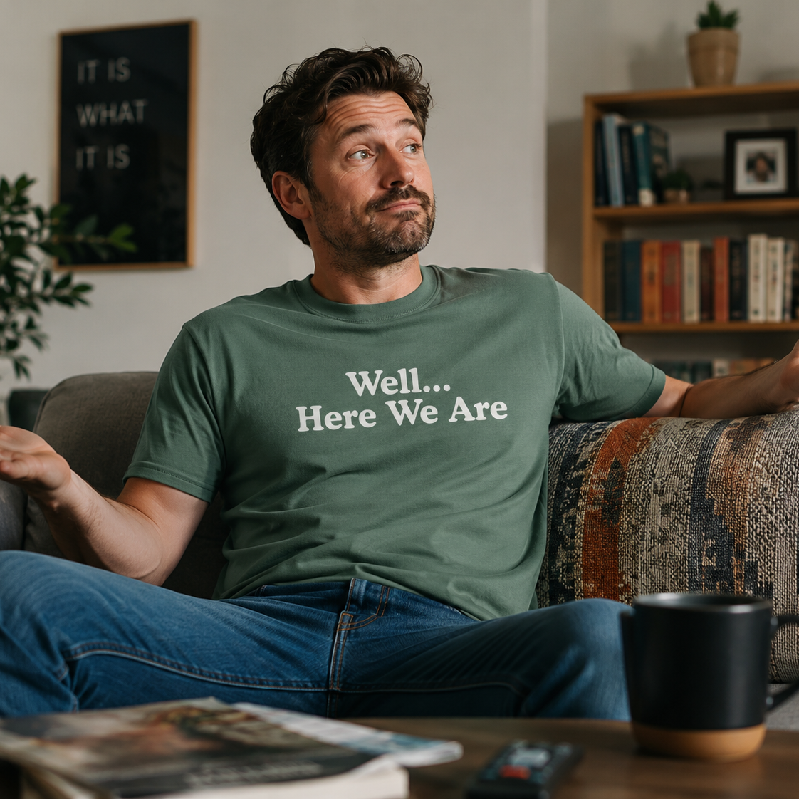 A man sitting on a couch in a living room wearing a green t-shirt with the white printed text 'Well... Here We Are'.