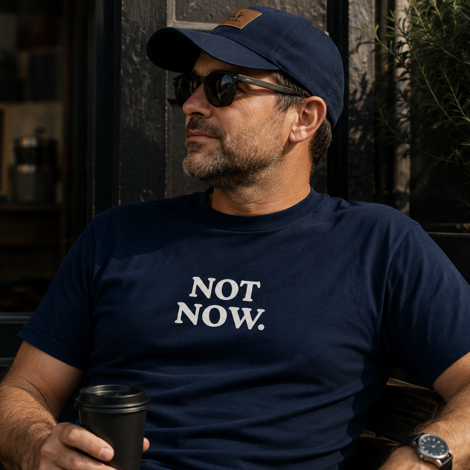 Man wearing a navy t-shirt with 'NOT NOW.' text, holding a coffee cup, sitting outdoors.