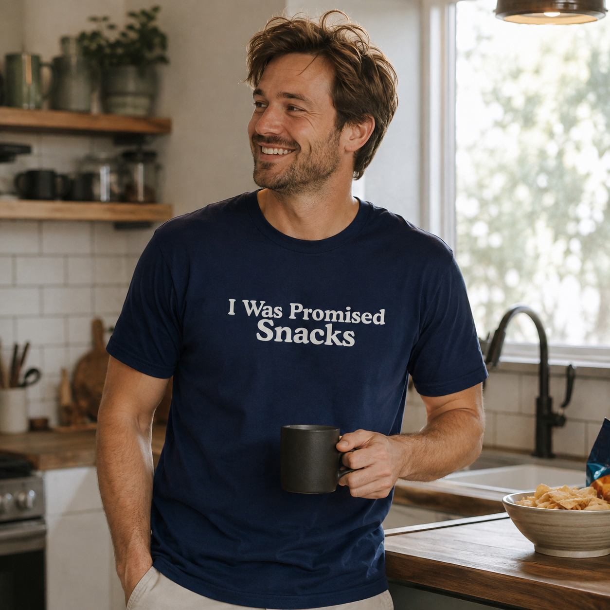 A man in a kitchen wearing a navy blue t-shirt with the white printed text 'I Was Promised Snacks'. He is holding a black mug and smiling, with a bowl of snacks on the counter in the background.