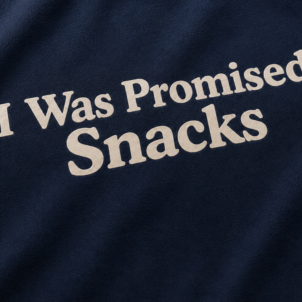 A close-up of a navy blue fabric surface with the phrase 'I Was Promised Snacks' printed in large cream-colored serif letters.
