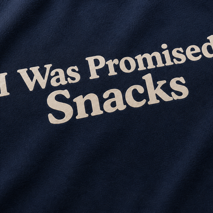A close-up of a navy blue fabric surface with the phrase 'I Was Promised Snacks' printed in large cream-colored serif letters.