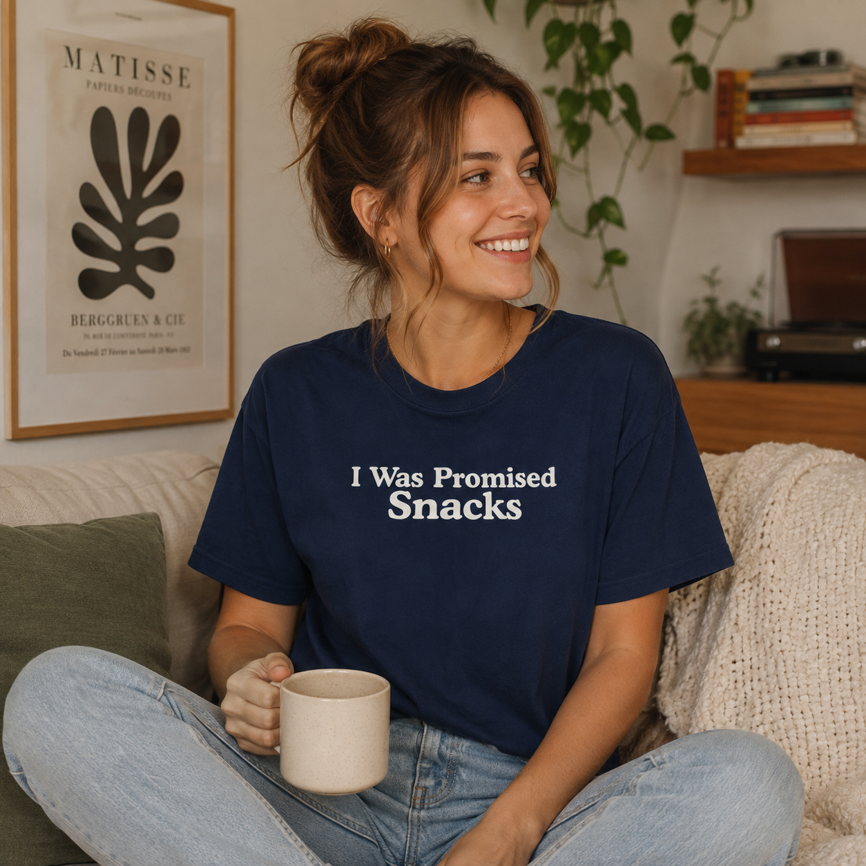 A woman sitting on a couch in a cozy living room setting, wearing a navy blue t-shirt with the white text 'I Was Promised Snacks' printed on the front. She is holding a beige ceramic mug.