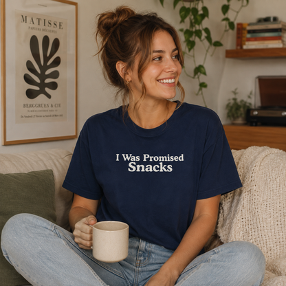 A woman sitting on a couch in a cozy living room setting, wearing a navy blue t-shirt with the white text 'I Was Promised Snacks' printed on the front. She is holding a beige ceramic mug.