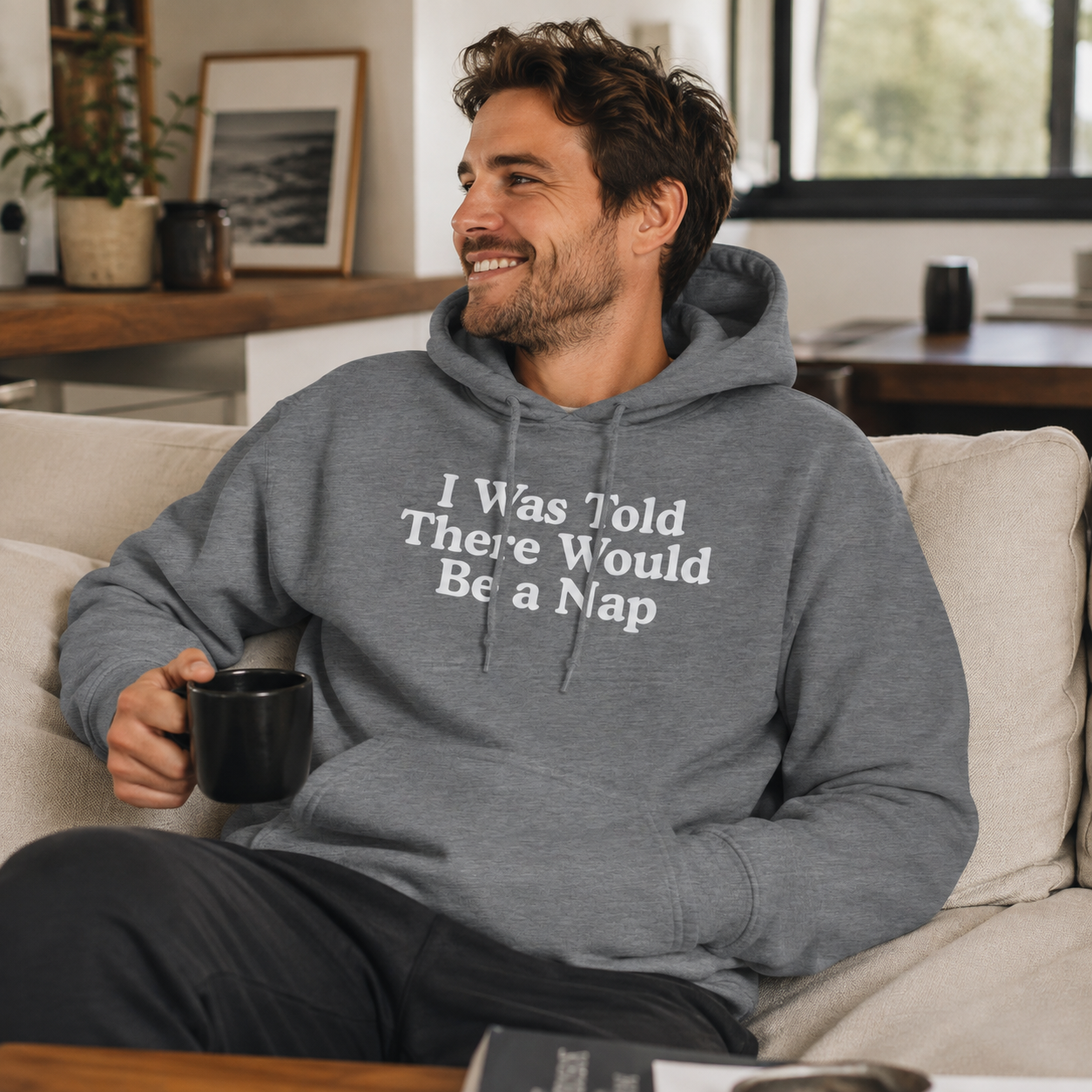 A man sitting on a beige couch in a living room, wearing a heather gray hoodie with the white text 'I Was Told There Would Be a Nap' across the chest. He is holding a black mug.