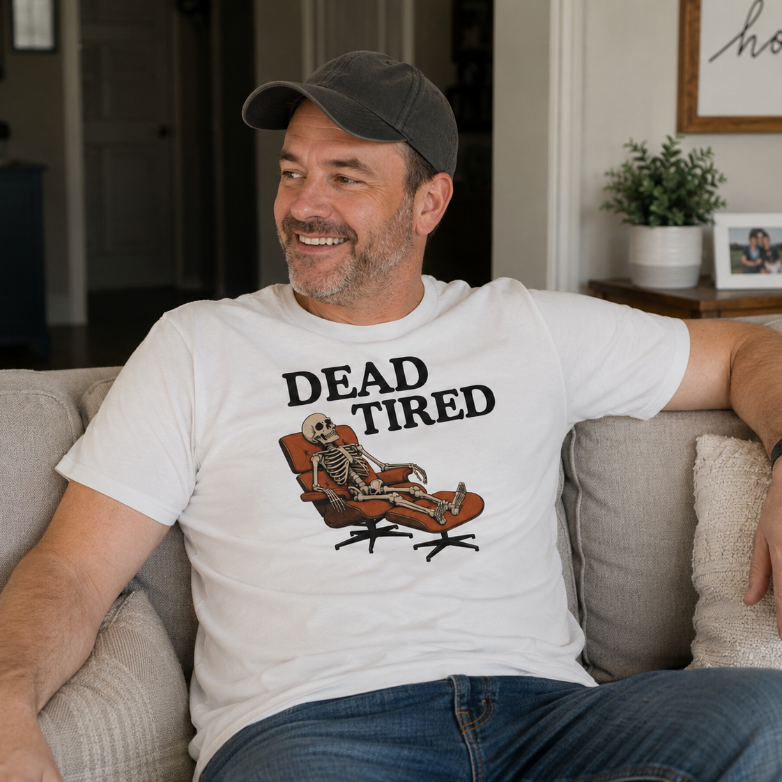 A man sitting on a beige couch wearing a white graphic t-shirt with the text 'DEAD TIRED' and a skeleton lounging in an orange chair, paired with a dark gray baseball cap and blue jeans.