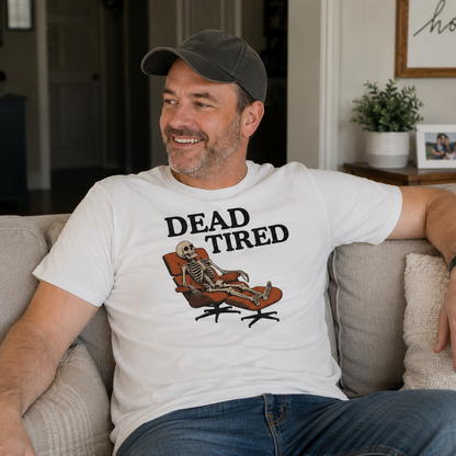 A man sitting on a beige couch wearing a white graphic t-shirt with the text 'DEAD TIRED' and a skeleton lounging in an orange chair, paired with a dark gray baseball cap and blue jeans.