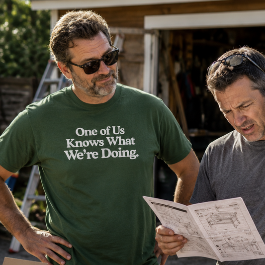 Two men standing outdoors at a worktable in a garage-like setting, with one man wearing a green graphic T-shirt that reads 'One of Us Knows What We're Doing.' and looking at a printed sheet.