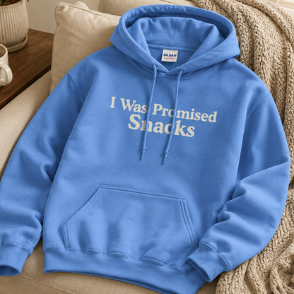 A blue pullover hoodie with the phrase 'I Was Promised Snacks' printed in white across the chest, styled on a beige couch with a knit throw blanket and a small plant nearby.