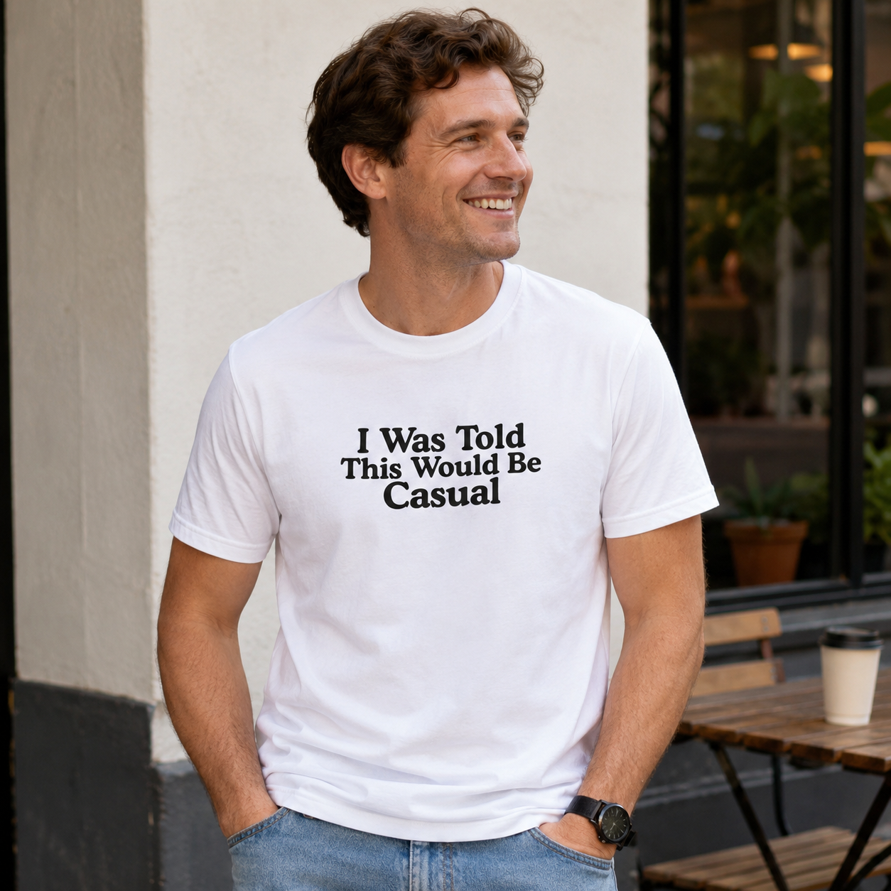 A man wearing a white crewneck t-shirt with the black printed text 'I Was Told This Would Be Casual' stands outdoors in front of a building wall.