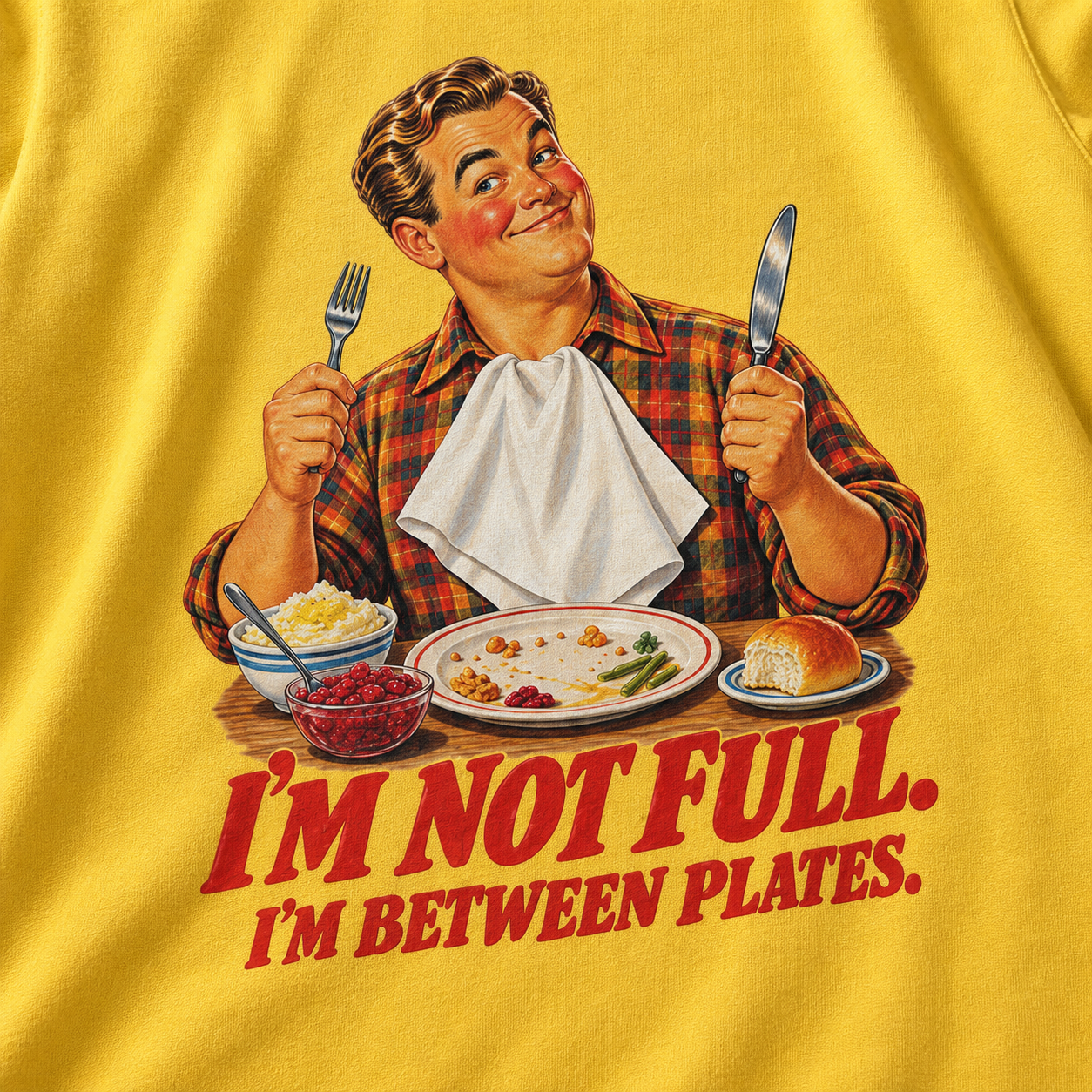 A close-up of a yellow graphic t-shirt featuring a retro illustration of a man holding a fork and knife, with plates of food and the text 'I'M NOT FULL. I'M BETWEEN PLATES.
