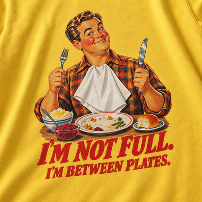 A close-up of a yellow graphic t-shirt featuring a retro illustration of a man holding a fork and knife, with plates of food and the text 'I'M NOT FULL. I'M BETWEEN PLATES.