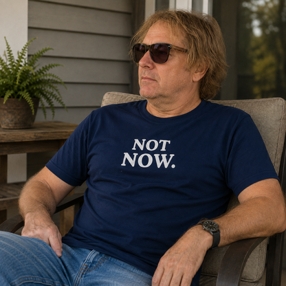 A man wearing a navy blue t-shirt with the text 'NOT NOW.' printed in white across the chest, sitting in a patio chair outdoors.