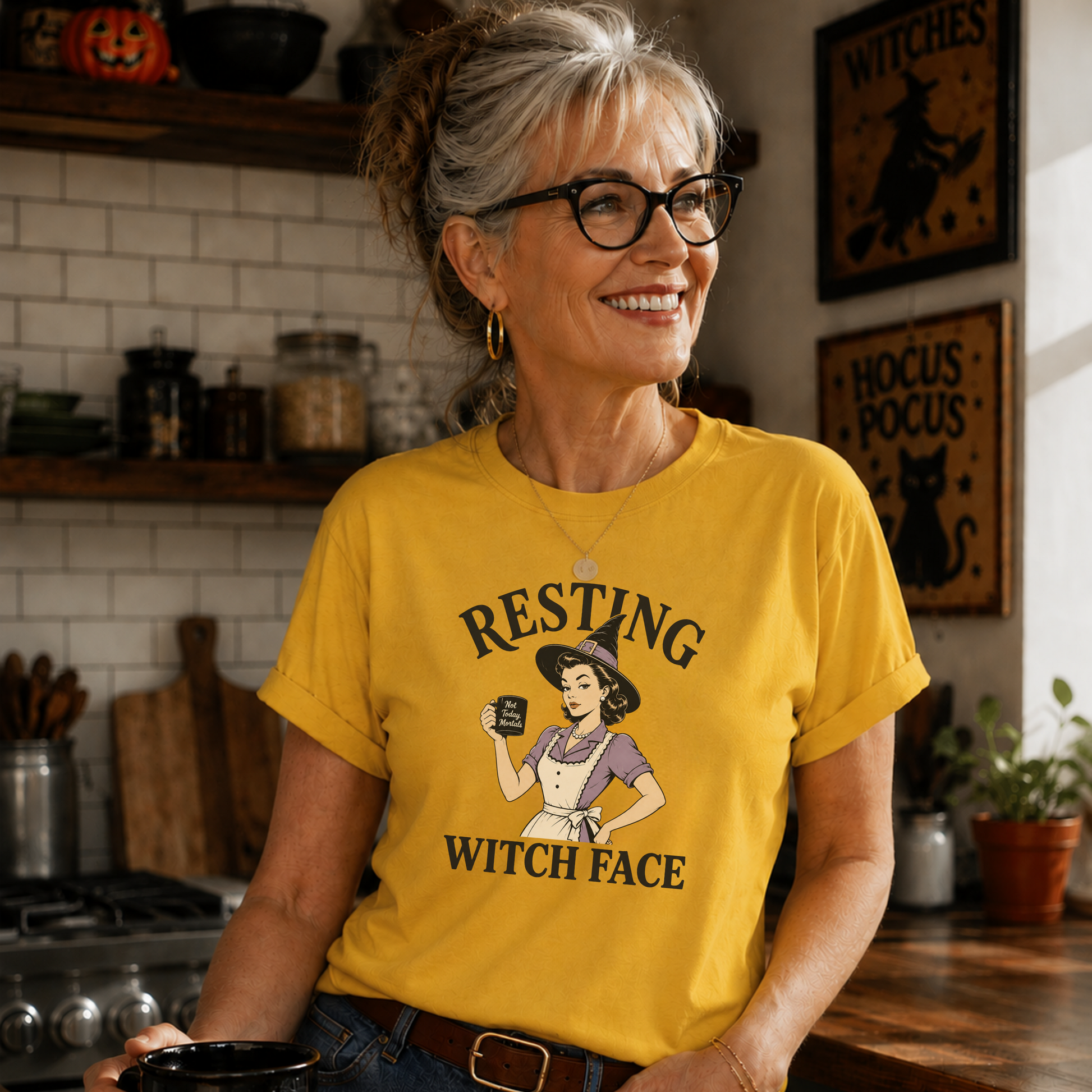 A smiling older woman with glasses stands in a cozy kitchen wearing a bright yellow graphic T-shirt that reads 'RESTING WITCH FACE' with an illustrated witch holding a mug. She is also wearing blue jeans and holding a black mug.