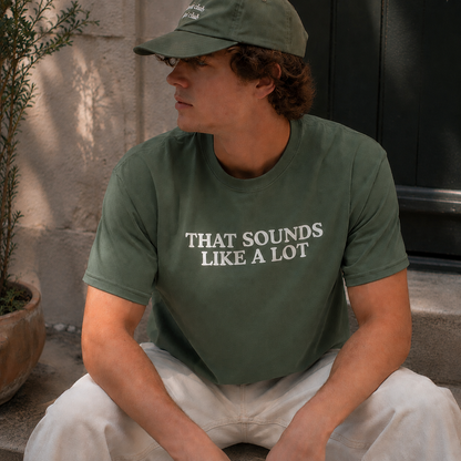A man wearing an olive green t-shirt with the text 'THAT SOUNDS LIKE A LOT' in white, paired with a matching olive green baseball cap and off-white pants, sitting outdoors.